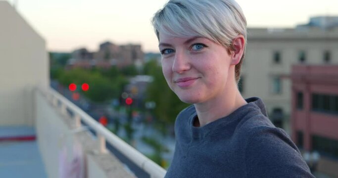 Happy flirtatious caucasian white girl looking into camera smiling from rooftop, then looks away with city street lights and buildings in background. In Cinema 4K (4096x2160) 30fps slowed from 60fps.