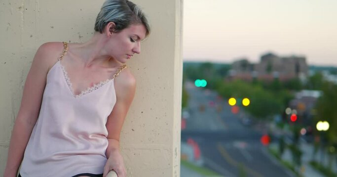 Short haired blonde girl with dark roots looking happy, then peeking over edge of tall building onto the city street at dusk, then back at camera. In Cinema 4K (4096x2160) 30fps slowed from 60fps.