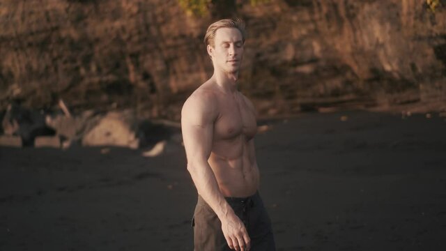 Athletic Caucasian man walking topless on the seashore after workout. Front view of young sportsman walking by wet sand on beach with elastic band. Man's beauty concept. High quality 4k footage