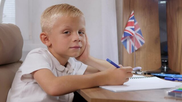 Smiling boy sitting at desk with notebooks and textbooks, doing homework and looking at camera over room interior background. Education online, children classes and studying concept.