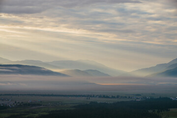 Scenic mountain landscape with orange lilac low clouds above village among mountains silhouettes under dawn cloudy sky. Atmospheric alpine scenery of countryside in low clouds in sundown golden color.