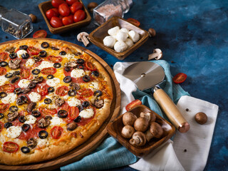 Beautiful composition. On a blue background, a large pizza on a wooden dish and many ingredients, a...