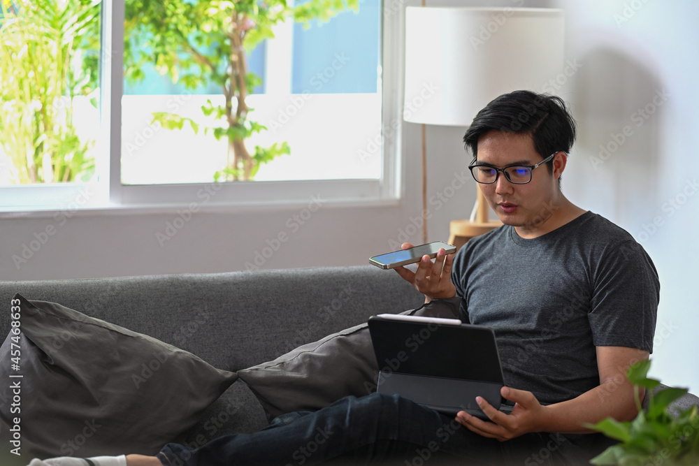 Poster Handsome man talking on speaker phone and using computer tablet on sofa.