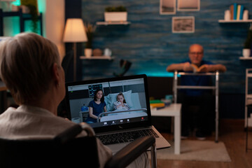 Aged woman with handicap using video call with laptop at home. Senior person talking to daughter and child while sitting in wheelchair in living room with online remote conference