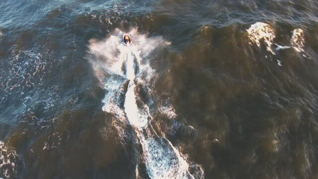 Aerial view of Young man rides on fast water scooter jumping on shallow waves and making golden splashes on sunset in blue water. Drone photo of tourist having fun riding a water bike on the sea