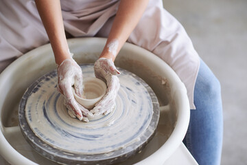 Hands of young woman working on pottery wheel in cozy workshop and making vase or mug
