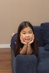 Cute female face portrait of little smiling girl, pretty adorable asian child lying on blue couch...