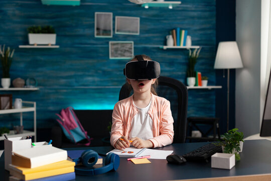 Little schoolchild wearing virtual reality headset enjoying playing with vr glasses sitting at desk in living room. Child studying literature doing school homework. Home schooling, distance education