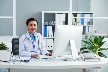 Portrait of smiling general practitioner in labcoat working on computer at her office desk