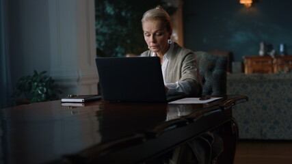 Rich mature woman typing laptop home. Greyhaired lady working computer cabinet.
