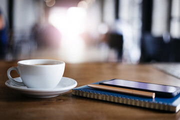 Closeup white cup of coffee with smartphone, notebook and pen on table in cafe. Vintage light, blurred and bokeh background