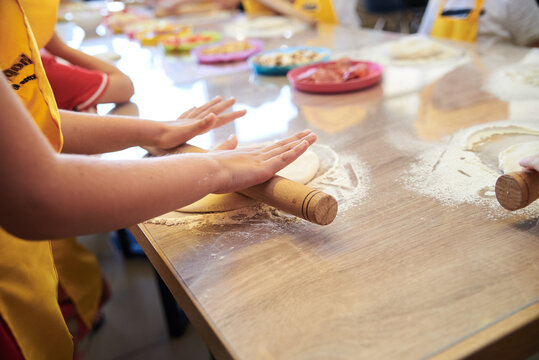 Child wearing yellow apron, making dough on the table. Close-up picture of hands, stretching dough with rolling pin. Bakery master class for small children. Pizza workshop at juniors' party.