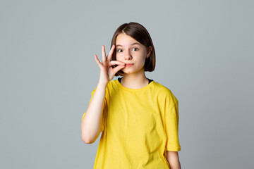 Portrait of a pretty teen girl showing zip gesture as if shutting mouth on key, standing over light grey background. You must be silent. Ban. Taboo. Censorship and harassment of freedom of speech - obrazy, fototapety, plakaty