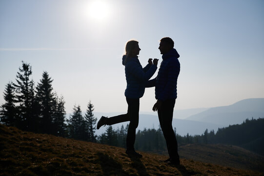 Silhouettes of girl standing on lawn with two arms outstretched to her boyfriend with one bent knee and raised leg, and man standing against girl supporting her by hand of backdrop of mountains afar.