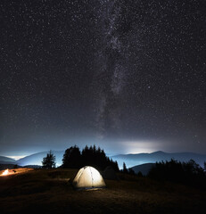 Fototapeta na wymiar Fantastic view of night starry sky over grassy hill with illuminated tourist tent. Magnificent scenery of mountain valley with camp tent under night sky with stars and Milky Way. Concept of camping.
