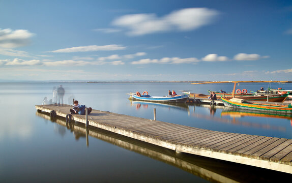 Calm sea with a wooden pier, boats, people and sky reflected in the water