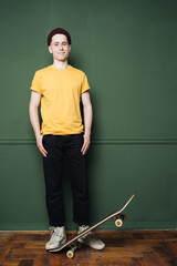 Portrait of young fashionable hipster man posing over green wall background. Caucasian male holding skateboard and smiling