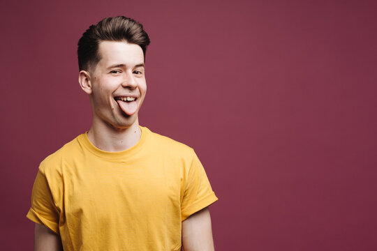 Emotional Young Handsome Brunette Man Student Wearing Yellow T Shirt Grimacing, Making Mouths, Sticking Out His Tongue At Camera Trying To Tease Someone, Acting Like A Little Naughty Child.