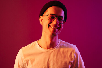 Shot of young handsome male with glasses trendy hairdo, wears casual white t shirt, has positive expression, poses in studio against pink background splashed by colorful lights