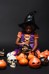 african american girl in witch costume and pointed hat looking at carved pumpkin on black