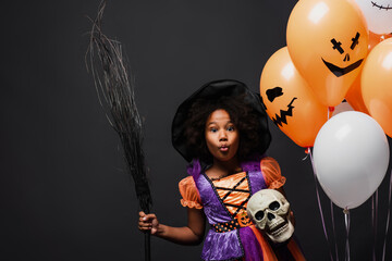 african american girl in halloween witch costume holding broom and skull near balloons isolated on...