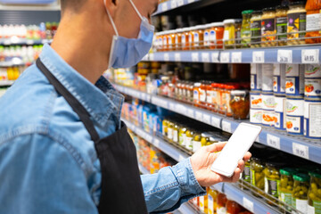Shopkeeper wearing a mask using smart phone to check the prices of the products at grocery store during virus epidemic outbreak.