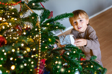 Portrait of a toddler boy wearing pajamas decorating a Christmas Tree at home. Winter holiday concept. Child waiting for a wonder.