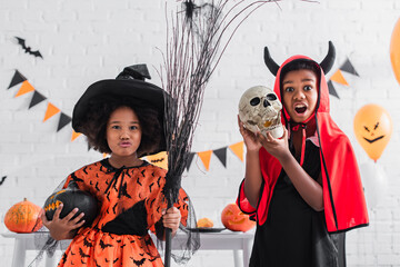 creepy african american boy in halloween costume holding skull near spooky sister with broom and...