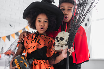 spooky african american kids in halloween costumes holding skull, carved pumpkin and broom