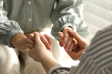 Religious women holding hands and praying together near window indoors, closeup