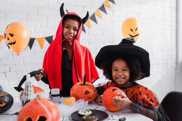 happy african american children in halloween costumes near carved pumpkins and cookies on table