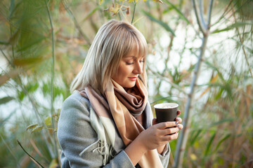 Blonde woman with short haircut in gray wool coat and checkered scarf drinks coffee cappuccino with marshmallows.