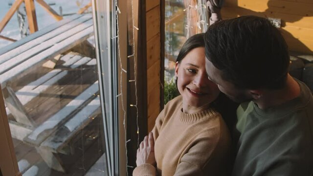 Handheld high angle slowmo shot of cheerful man walking up to his wife standing by window in cabin and enjoying winter day. He is hugging her and smiling affectionately
