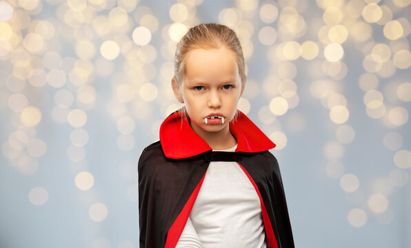 Halloween, Holiday And Childhood Concept - Girl In Black Dracula Costume With Cape And Fangs Over Festive Lights On Grey Background