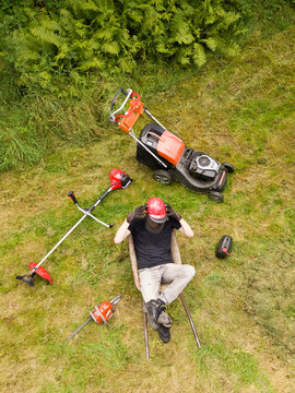 Gardener resting and sitting in a wheelbarrow tired after mowing the grass and cutting branches. Around it is a mower, a brushcutter and a chainsaw. View from above