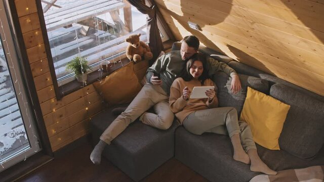 High angle shot of cheerful man and woman relaxing on couch in cozy cabin and browsing internet on mobile phone and tablet