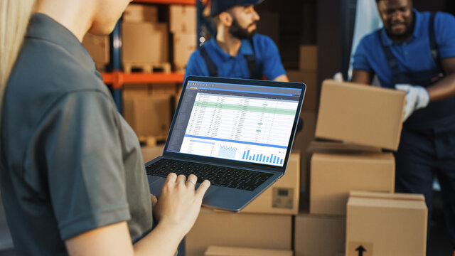 Female Manager Using Laptop Computer To Check Inventory. In The Background Warehouse Retail Center With Cardboard Boxes, E-Commerce Online Orders, Food, Medicine, Products Supply. Over The Shoulder