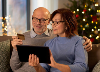 online shopping, winter holidays and people concept - happy smiling senior couple with tablet pc computer and credit card at home in evening over christmas tree lights on background