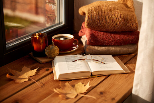 season and objects concept - open book with glasses, cup of coffee, autumn leaves, wool sweaters and candle burning on wooden window sill at home