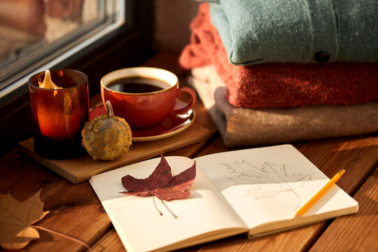 season and objects concept - open sketchbook with drawing of autumn leaf, cup of coffee, wool sweaters and pumpkin on wooden window sill at home