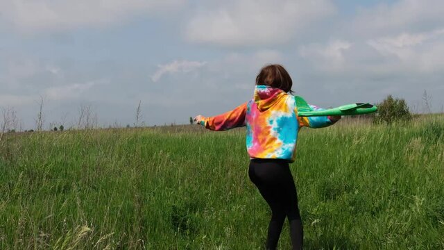 Beautiful girl wearing colorful hoodie playing with toy plane in the field with green grass and smiling. Pretty female teenager launching airplane at the nature and feel freedom