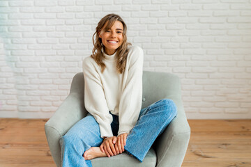 Indooor winter portrait of pretty woman in cozy sweater sittinh on modern sofa at home.