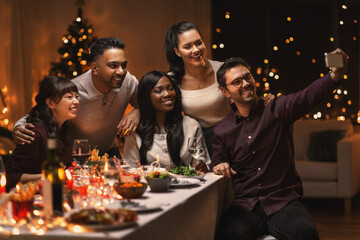 holidays, party and celebration concept - multiethnic group of happy friends having christmas dinner at home and taking selfie with smartphone