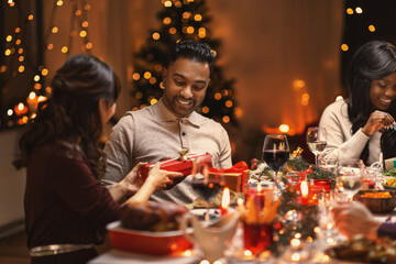 holidays, party and celebration concept - multiethnic group of happy friends having christmas dinner and giving presents at home
