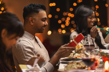 holidays, party and celebration concept - multiethnic group of happy friends having christmas dinner and giving presents at home
