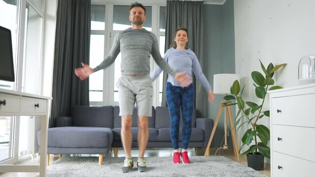 Caucasian couple is doing jumping jacks exercise at home in cozy bright room.