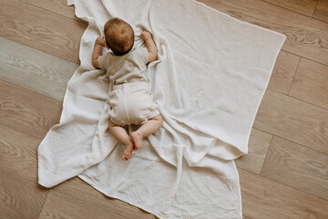 Baby lying on tummy on a white muslin blanket on the floor, top view. - obrazy, fototapety, plakaty