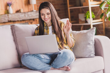 Photo of sociable relaxed lady sit cozy couch hold netbook wave hand toothy smile wear plaid shirt home indoors