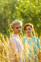 Lesbian young red haired woman stands by loving girlfriend in high meadow grass against green trees at gentle sunset light closeup - 457526007