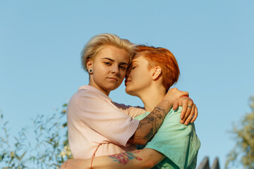 Lesbian attractive young women with stylish haircuts hug standing under clear blue sky at orange sunset light in summer evening closeup